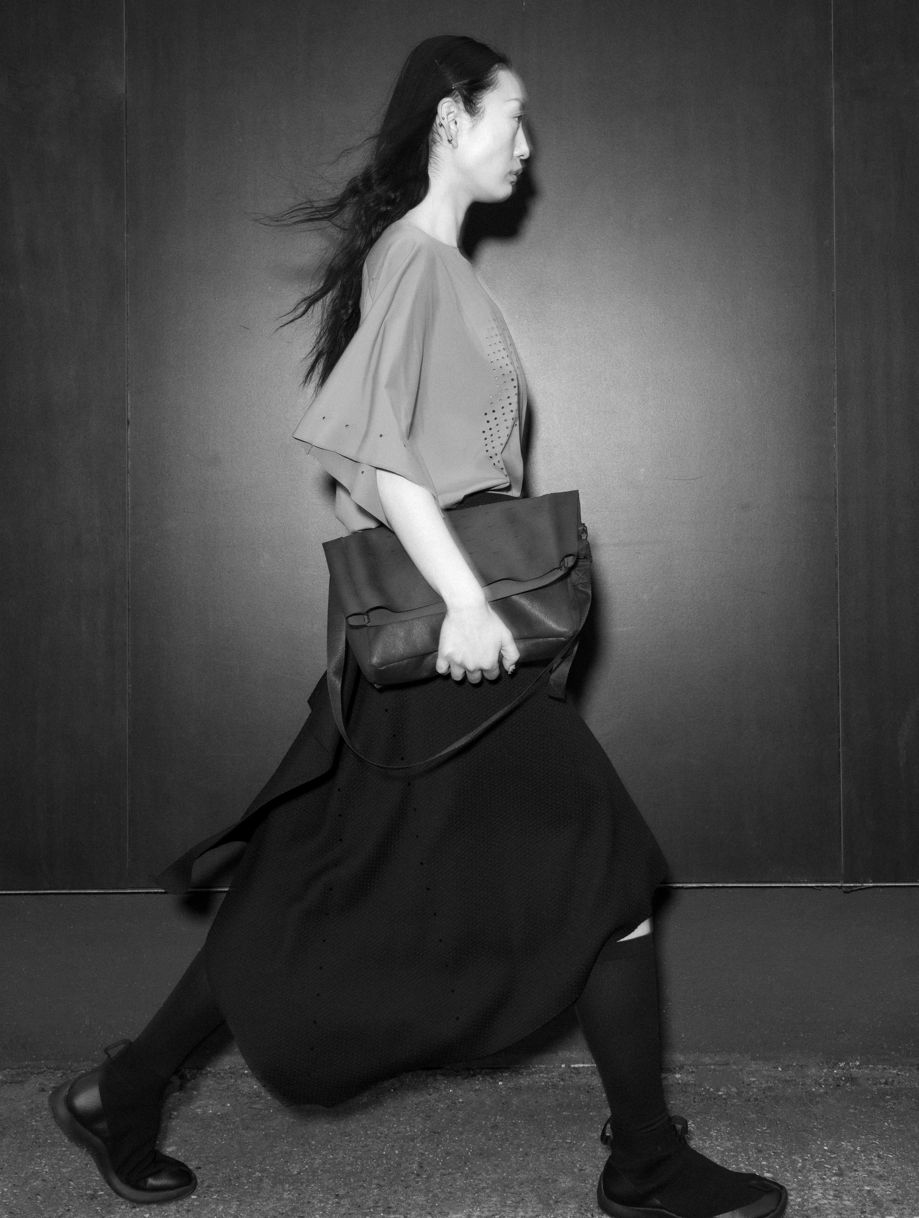 Woman wears fluid black skirt in motion, with short sleeve grey kimono t-shirt and leather folded handbag, by Savvas Alexander.